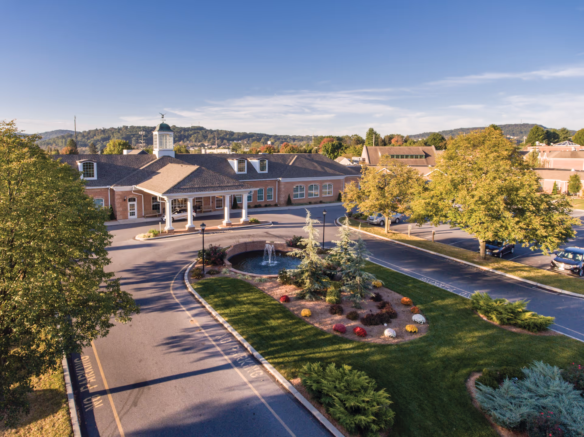 Aerial view of a senior living facility's front entrance with a circular driveway, fountain, and landscaped grounds.