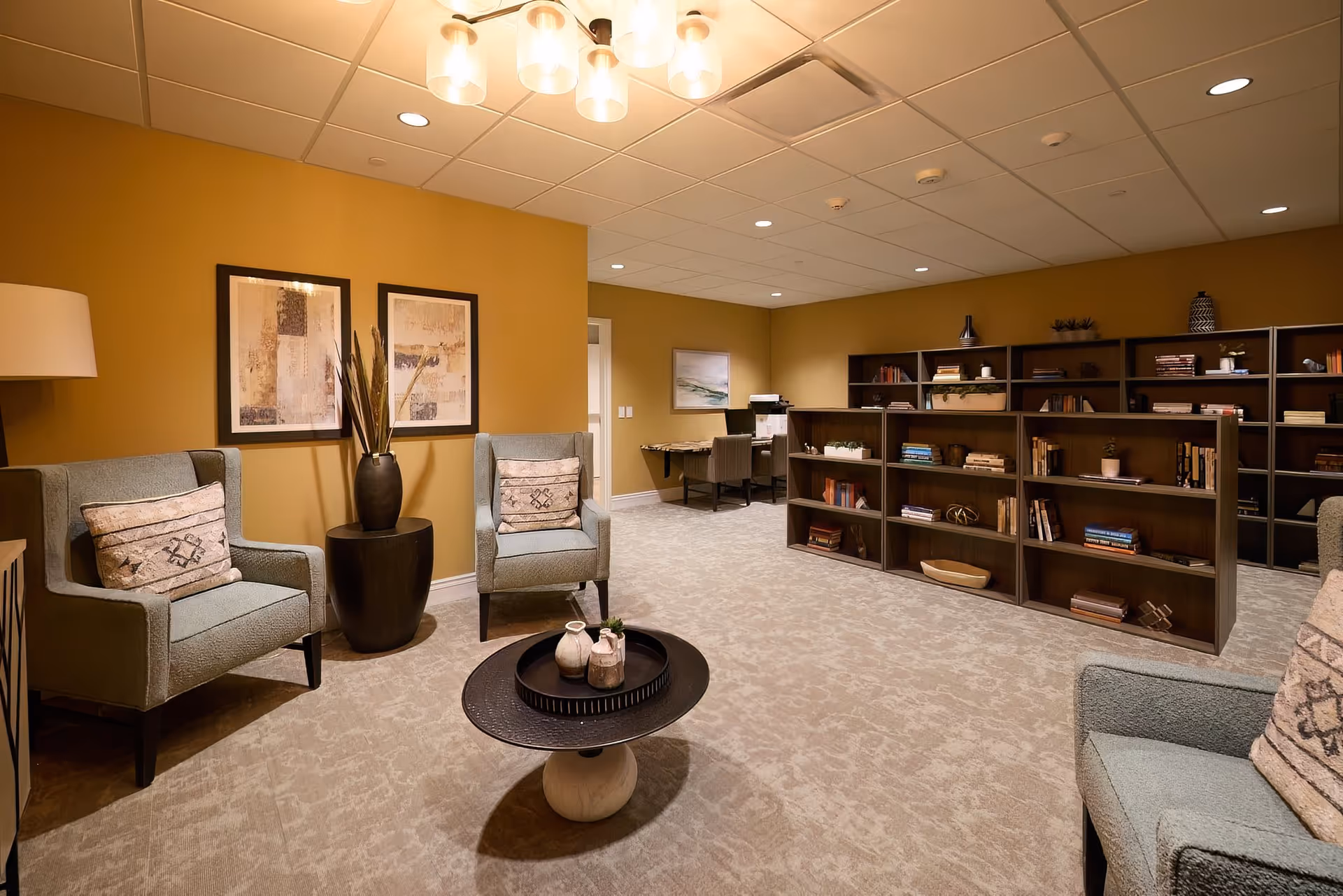 A cozy living room area with mustard yellow walls, two light blue armchairs with patterned cushions, a round black coffee table with decorative items, and a dark wooden bookshelf filled with books and decor. In the background, there is a small desk with a chair and a printer, and two framed abstract artworks on the wall.