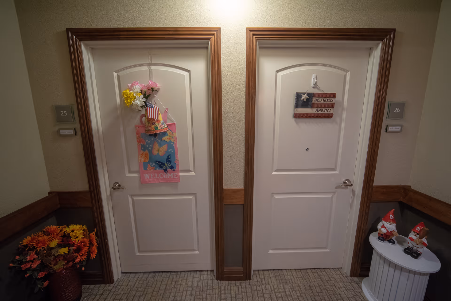Two white doors in a hallway of a senior living facility, each decorated with hanging signs. The left door has a colorful welcome sign with flowers and a small American flag, while the right door has a sign with the American flag and the words 'God Bless America'. There is a vase with artificial flowers on the floor to the left and a small white table with two gnome figurines to the right. The doors are numbered 25 and 26.