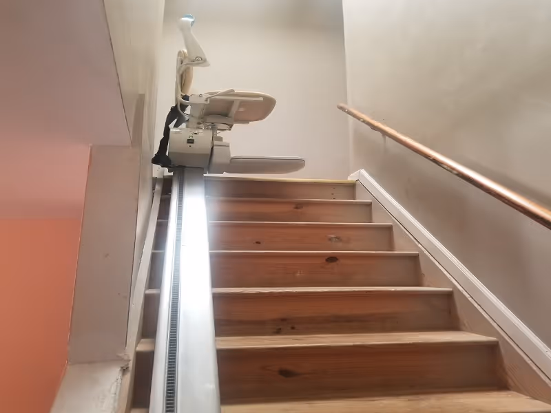 Wooden staircase with a stairlift chair installed on a rail alongside a wall handrail.