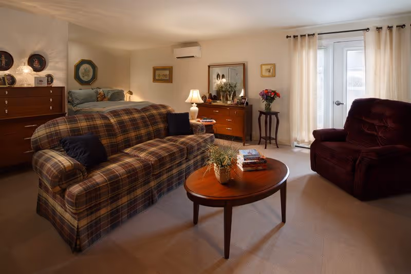 A cozy living area with a plaid sofa, a wooden coffee table with a plant and books, a burgundy armchair, and a dresser with a lamp and decorative items. Behind the sofa, there is a bed with pillows and a teddy bear, framed pictures on the walls, and a glass door with light curtains letting in natural light.