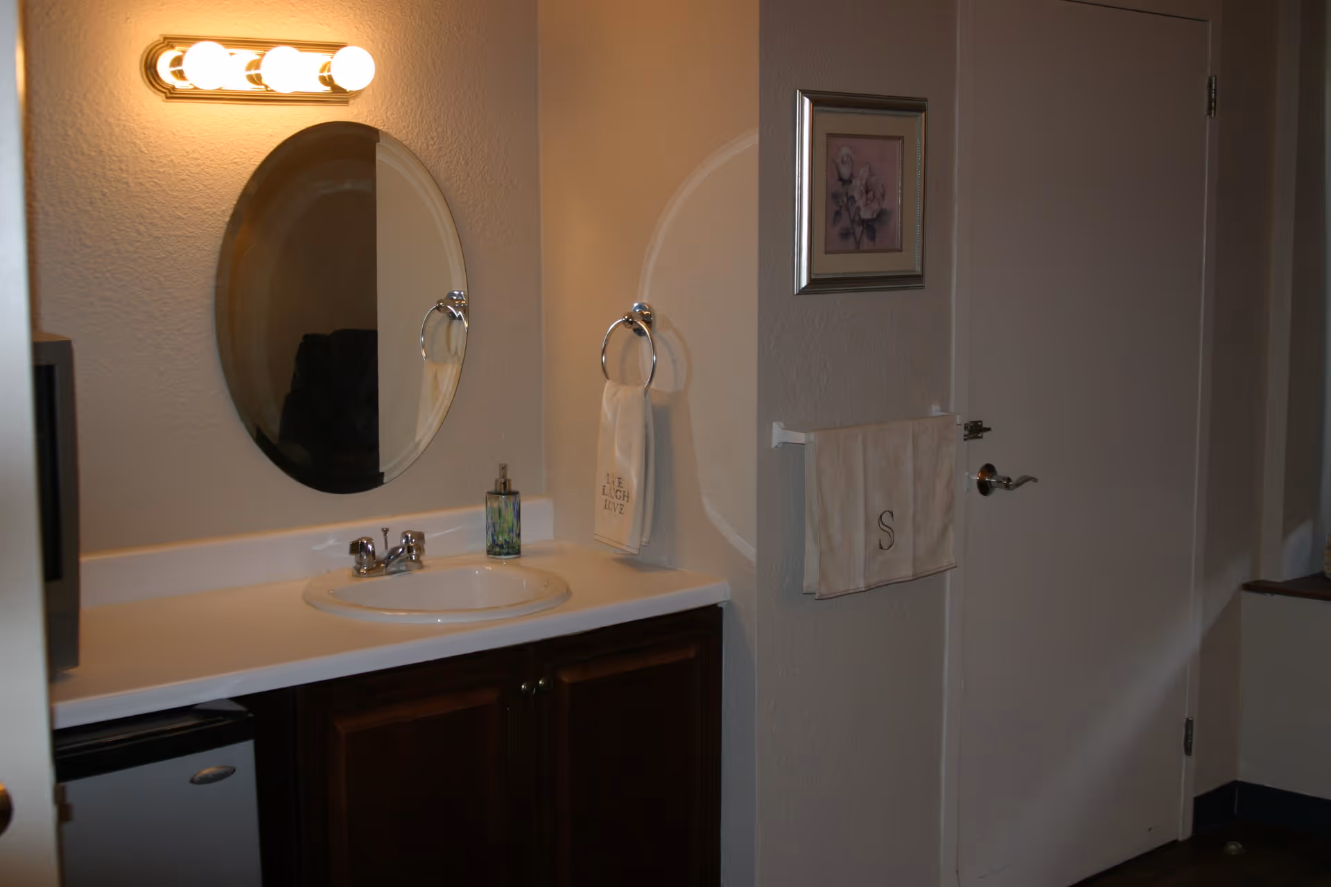 A bathroom vanity area with a white countertop, a round mirror mounted on the wall, and a light fixture with three bulbs above the mirror. There is a soap dispenser on the countertop and a towel ring with a white hand towel that says 'LIVE LAUGH LOVE'. On the adjacent wall, there is a framed floral picture and a towel rack with a beige towel monogrammed with the letter 'S'. A closed white door is visible next to the towel rack.