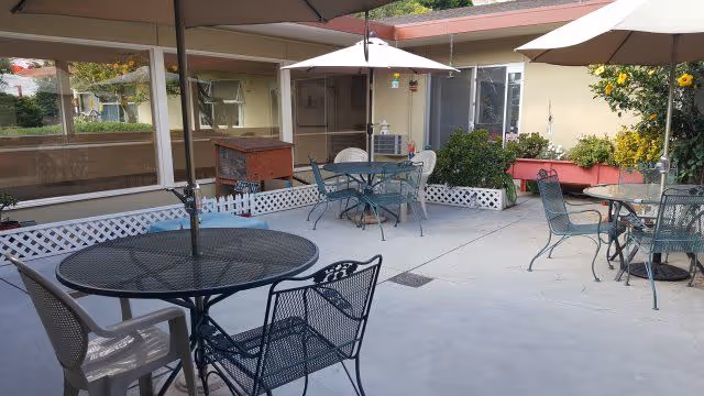Outdoor patio area with several metal tables and chairs, each table shaded by a large umbrella. The patio is surrounded by a low white lattice fence and some greenery, including bushes and flowers. The building walls and windows are visible in the background.