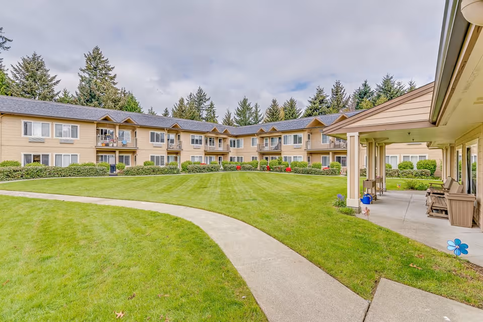 A well-maintained outdoor courtyard area of a senior living facility with a curved concrete walkway, green grass, shrubs, and a two-story beige building with balconies and windows surrounding the courtyard. There is a covered patio area with chairs on the right side and tall trees in the background under a cloudy sky.