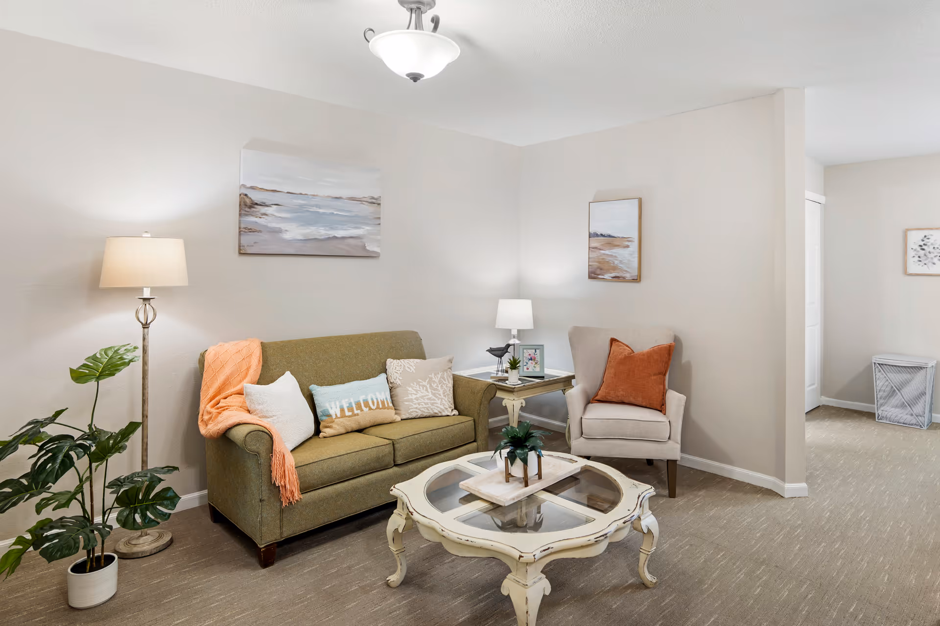 A cozy living room with a green sofa adorned with multiple pillows and an orange throw blanket. Next to the sofa is a floor lamp and a potted plant. There is a beige armchair with an orange pillow beside a small side table holding a lamp and framed photo. A decorative glass-top coffee table with a small plant centerpiece sits in front of the sofa. The walls are light-colored and decorated with two landscape paintings. The room has carpeted flooring and an open doorway leading to another area.