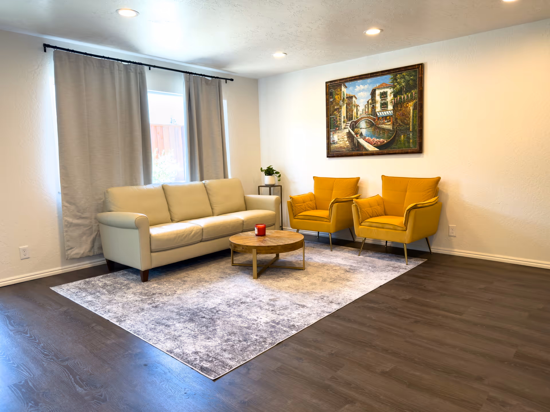 Bright living room with a beige sofa, two yellow armchairs, a round coffee table on an area rug, and a framed painting on the wall.