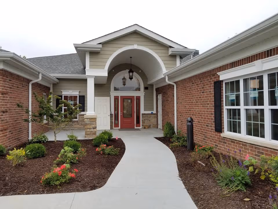 Front entrance of a senior living facility with a covered porch, red double doors, brick walls, and landscaped garden beds with shrubs and flowers along a curved concrete walkway.
