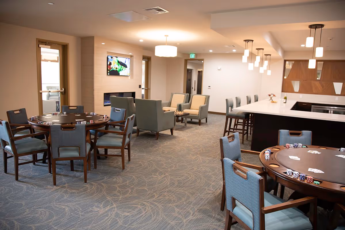 A spacious senior living common area featuring two round poker tables with chairs, poker chips, and playing cards. In the background, there is a seating area with armchairs and a small table with a chessboard. A wall-mounted TV and a modern fireplace are visible. To the right, there is a kitchen island with bar stools and pendant lights hanging above.
