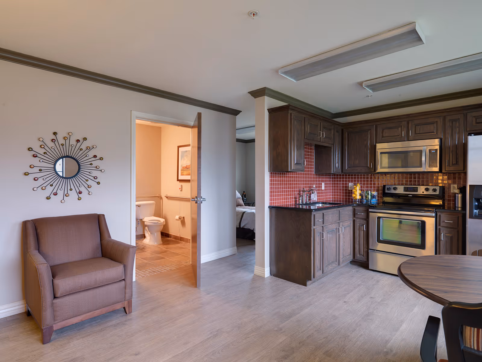 Open-plan living area with a kitchenette featuring dark wood cabinets and stainless steel appliances, a brown armchair, and an open door showing a bathroom and bedroom.