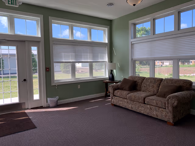 A bright room with light green walls featuring large windows with white blinds partially open, allowing sunlight to fill the space. There is a patterned sofa with three cushions against the wall, a small wooden side table with a lamp, a glass door with an exit sign above it, and a trash can near the door. The floor is carpeted in a neutral tone.