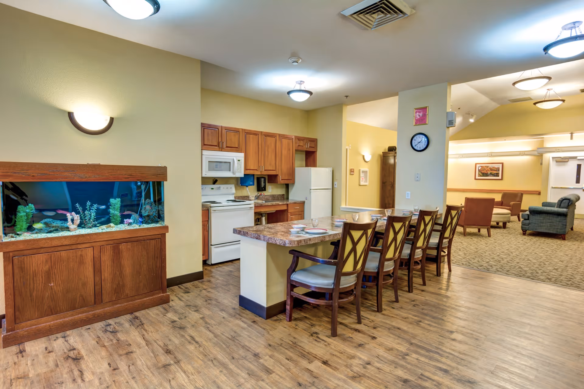 Interior view of a senior living facility showing a kitchen area with wooden cabinets, a white refrigerator, stove, and microwave. In front of the kitchen is a counter with six wooden chairs, each set with plates and glasses. To the left of the kitchen is a large fish tank with colorful fish and aquatic plants. The background shows a lounge area with armchairs and a carpeted floor, illuminated by ceiling lights.
