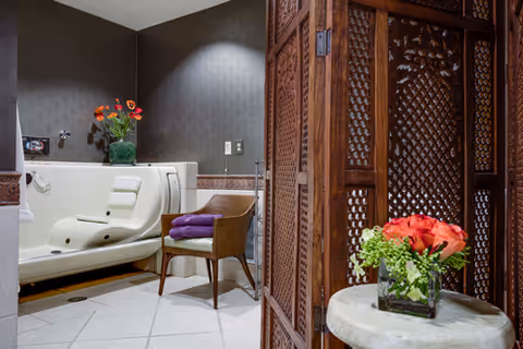A bathroom featuring a white walk-in bathtub with a built-in seat, a wicker chair with folded purple towels on it, and a vase of red flowers on a ledge. A decorative wooden folding screen partially obscures the view, and a small round table with a glass vase of red and green flowers is in the foreground.