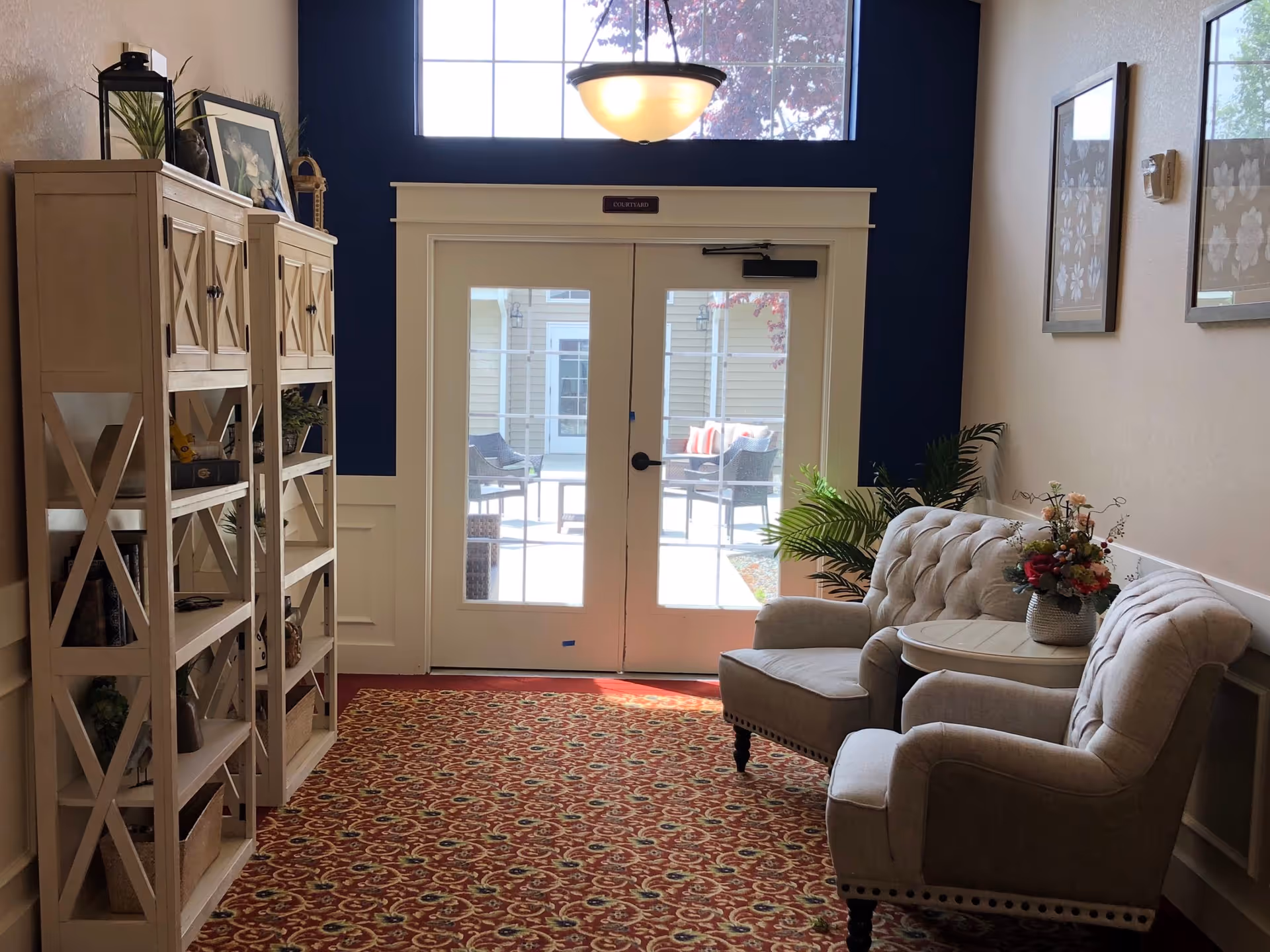 Sunlit seating area with two upholstered chairs, a small table with flowers, shelving, and double glass doors leading outside.
