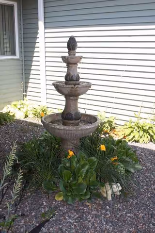 Stone tiered fountain surrounded by yellow flowers and green plants in front of a gray house.