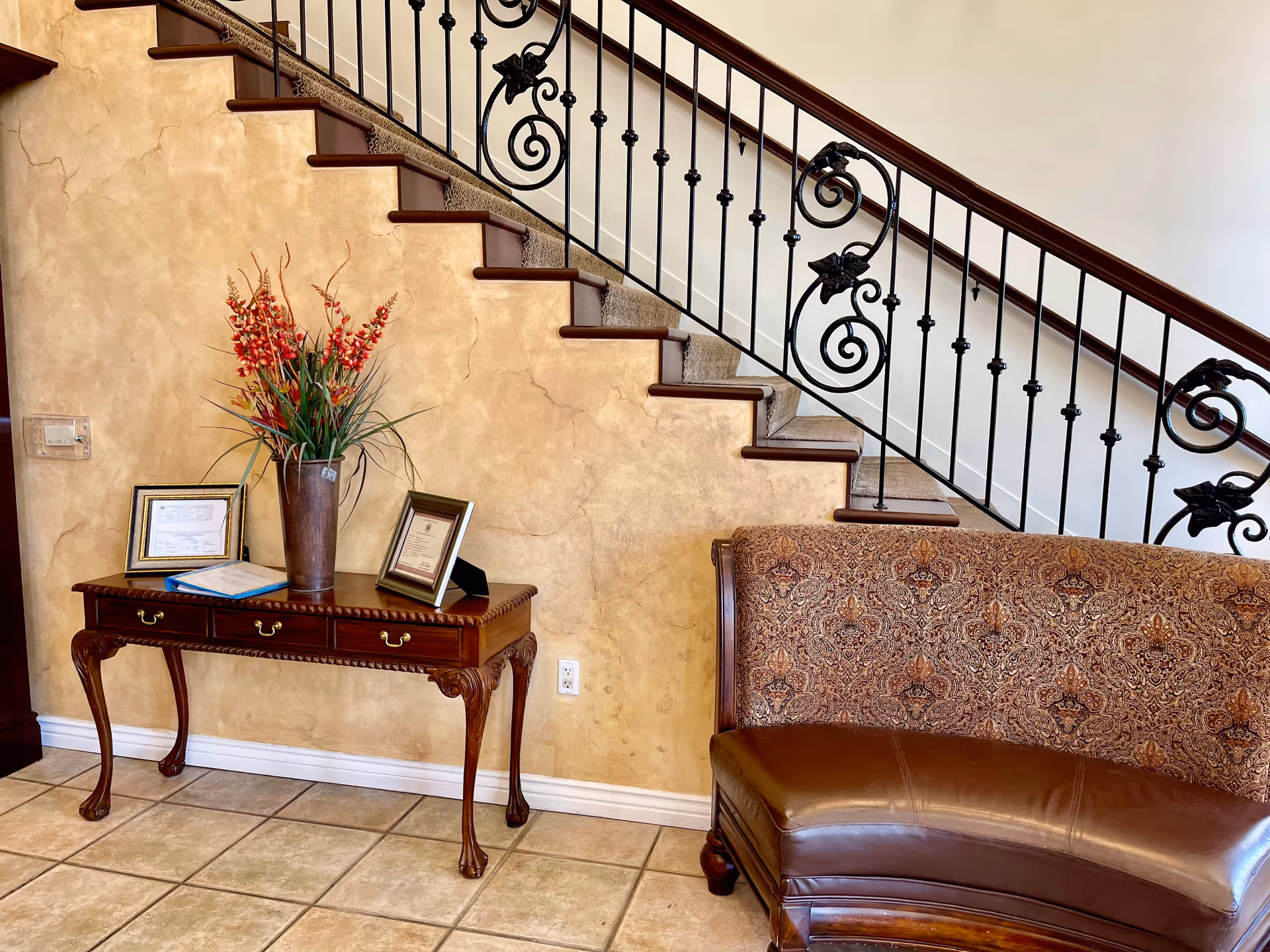 A decorative staircase overlooks a small console table with a vase of flowers and framed certificates next to an upholstered curved bench.
