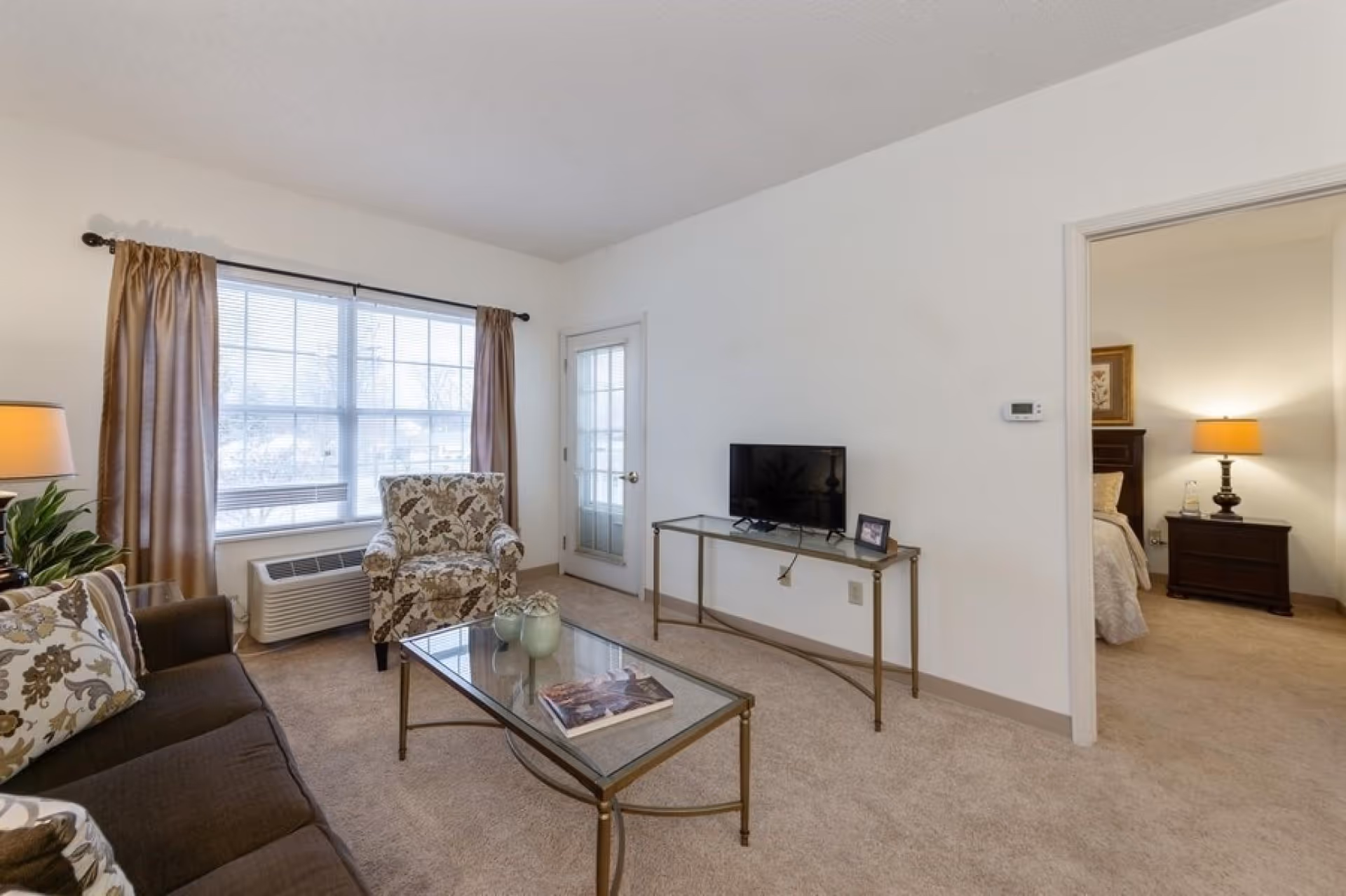 A cozy living room with a brown sofa adorned with patterned pillows, a floral armchair near a large window with beige curtains, a glass coffee table with decorative items, and a small TV on a glass console table. A doorway leads to a bedroom with a nightstand and lamp visible.