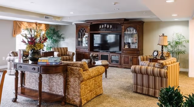 A spacious senior living common area with patterned carpet, a large wooden entertainment center with a flat-screen TV, and several upholstered armchairs and sofas arranged around a central coffee table. There is a wooden console table with books and a floral arrangement, and potted plants add greenery to the room.