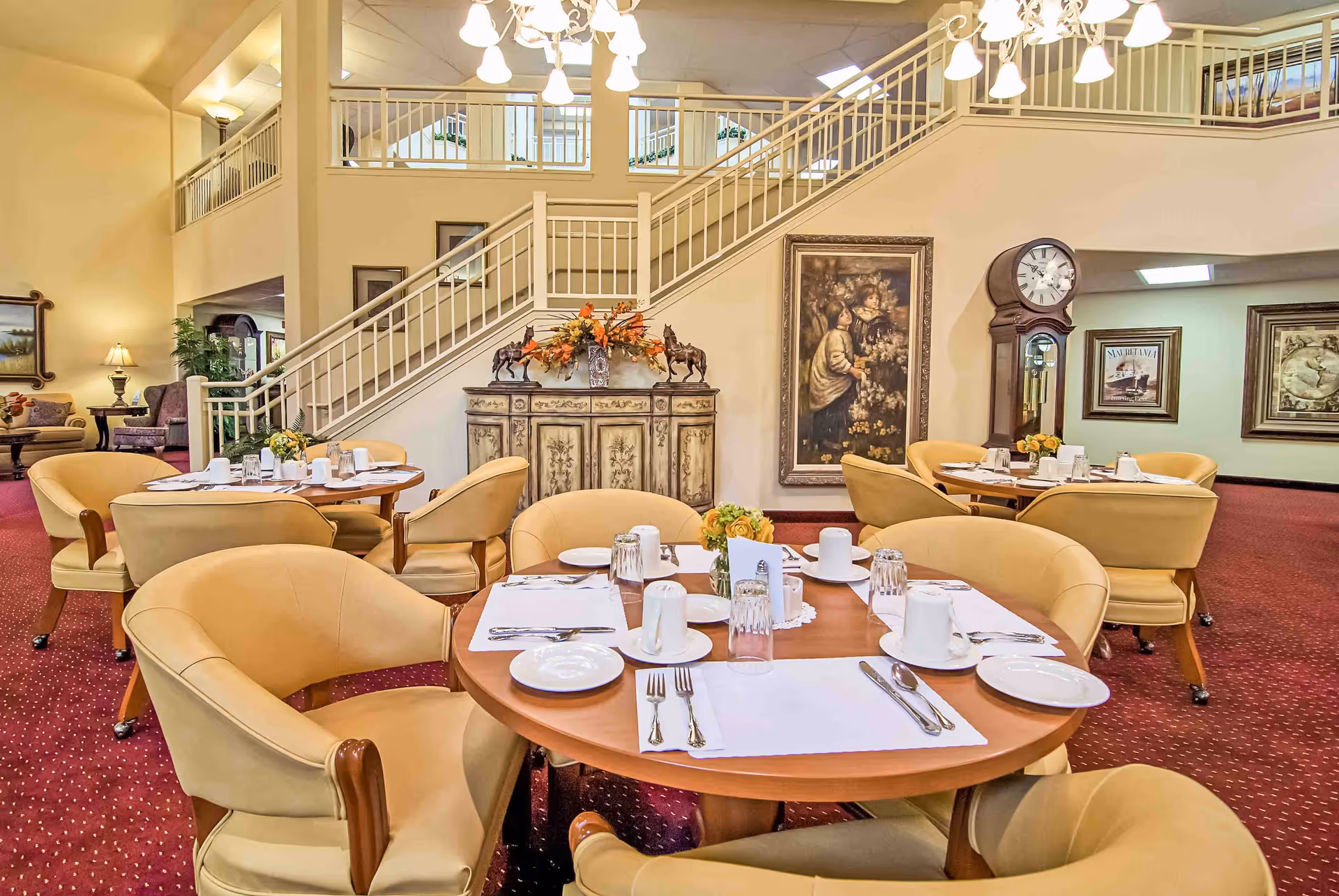 A bright dining room with round tables set for meals, beige upholstered chairs, a staircase, and decorative artwork and a grandfather clock.