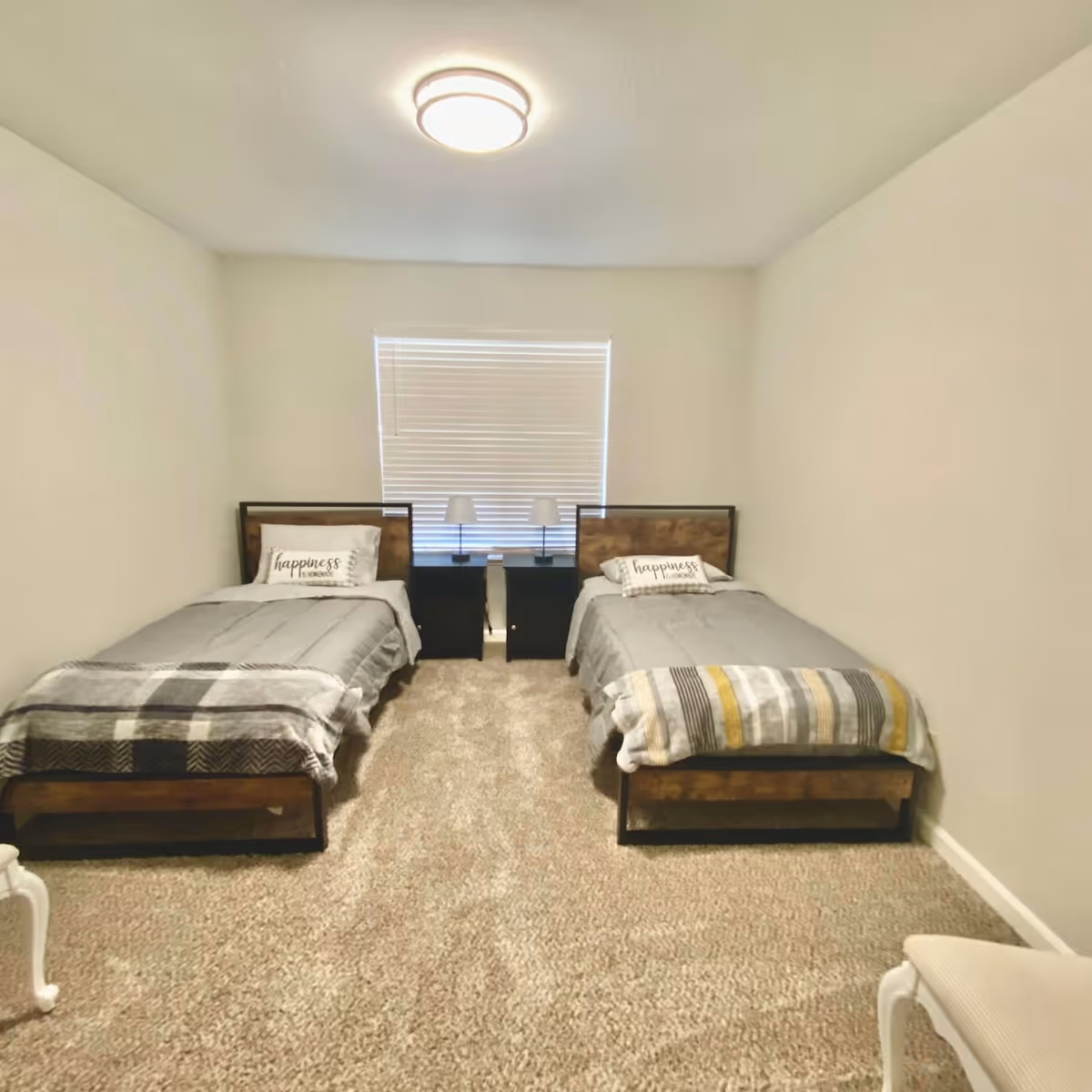 A simple bedroom with two single beds placed parallel to each other against opposite walls. Each bed has a gray blanket and a decorative pillow with the word 'happiness' on it. Between the beds is a window with closed blinds and two small black nightstands, each with a white lamp. The room has beige carpet flooring and plain white walls, with a ceiling light fixture above.