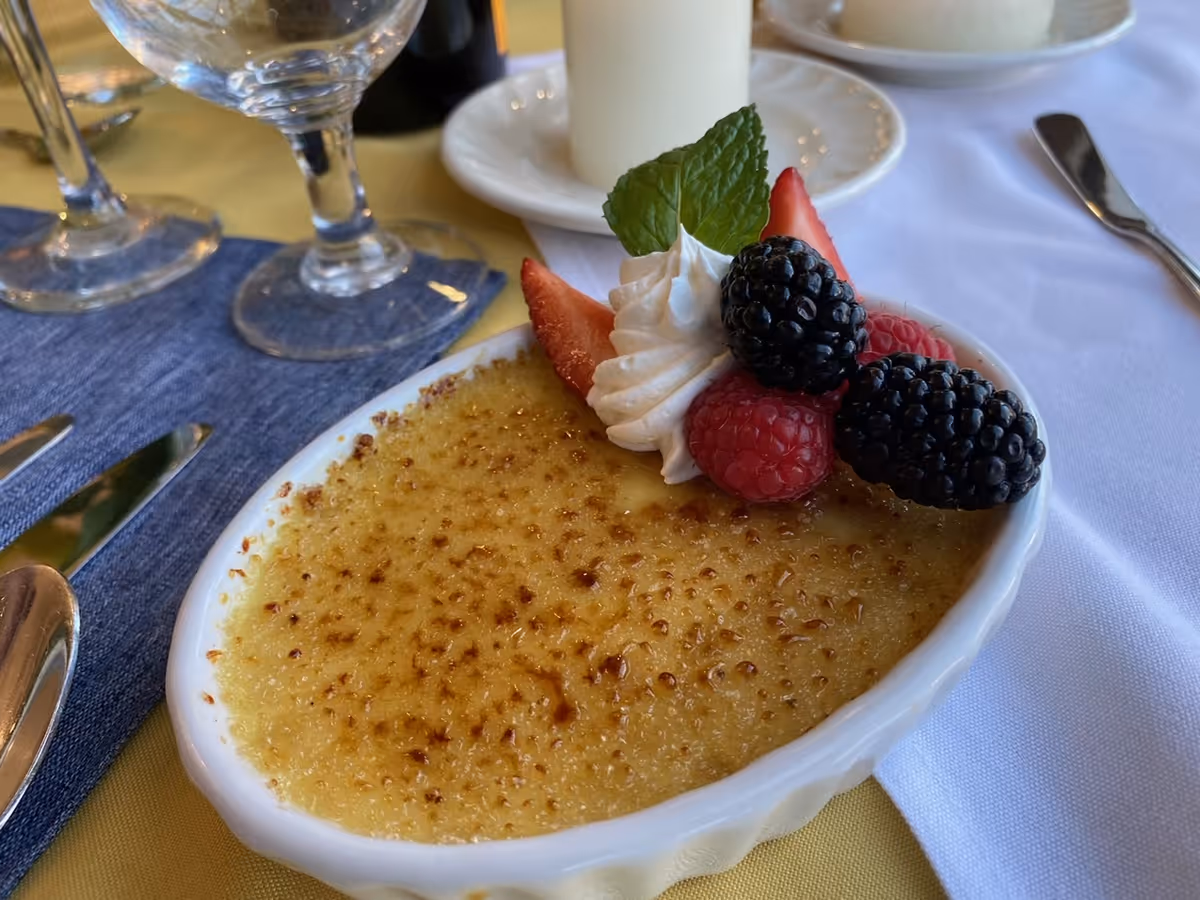 A close-up of a crème brûlée dessert topped with whipped cream, fresh strawberries, raspberries, and blackberries, served in a white oval dish on a table set with glassware, cutlery, and a white tablecloth.