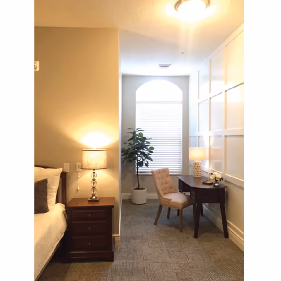 A cozy bedroom corner featuring a wooden nightstand with a lit lamp next to a bed with white bedding and a gray pillow. Adjacent to the bedroom area is a small workspace with a wooden desk, a beige upholstered chair, a table lamp, and a small flower arrangement. A large window with white blinds and an arched top is in the background, along with a potted plant. The walls are painted light beige with decorative paneling on one side.