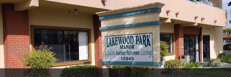 Exterior view of Lakewood Park Manor building with a sign that reads 'Lakewood Park Manor Quality Assisted Retirement Living 12045' surrounded by bushes and plants.