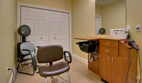 Interior view of a senior living facility's hair salon area featuring a salon chair, a hair washing station with a black sink, a wooden cabinet with drawers, a hair dryer mounted on the side, and a large mirror on the wall. There is also a small chair and a set of white double doors in the background.