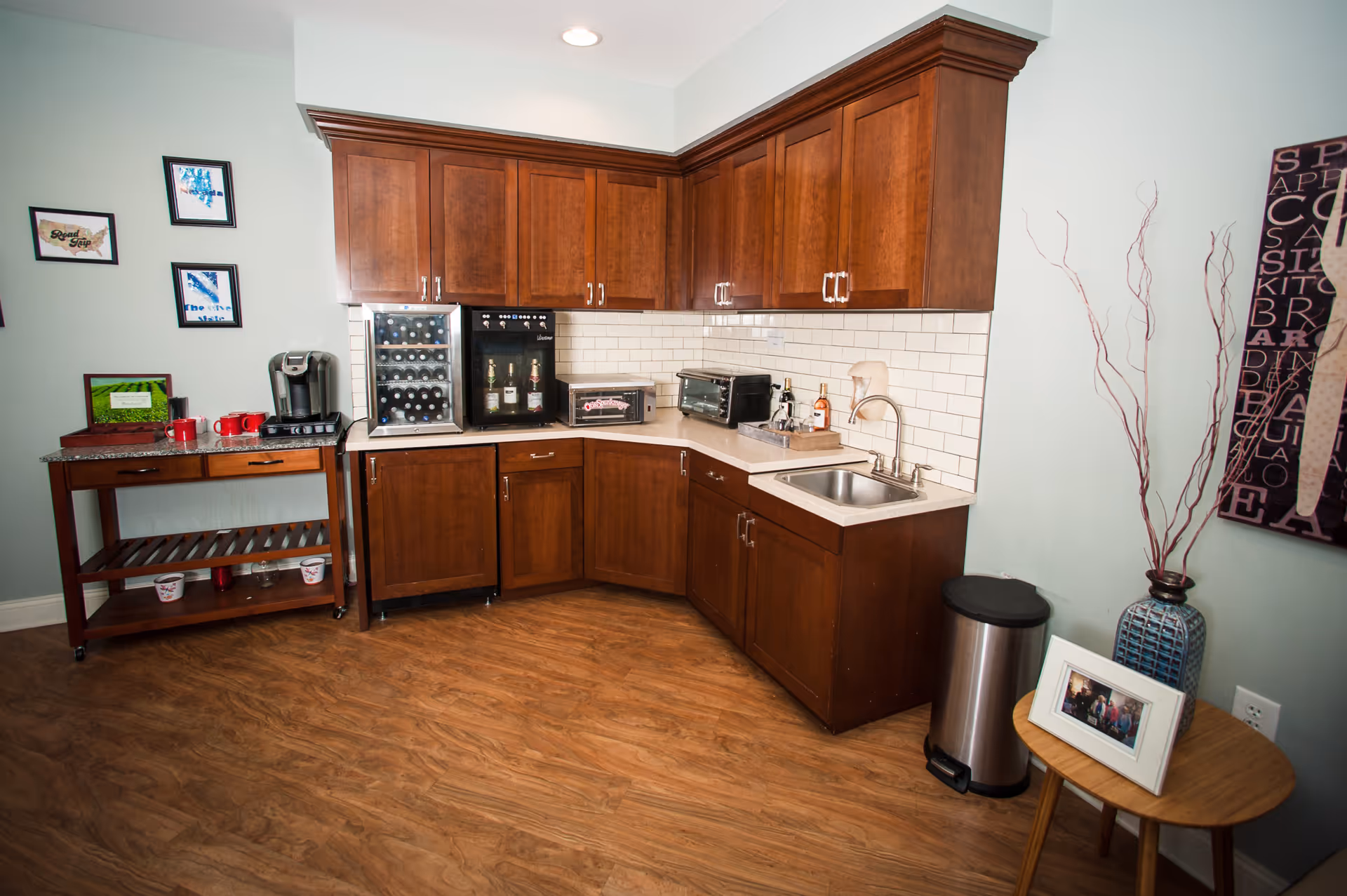 A small kitchenette area with wooden cabinets, a countertop with a sink, a wine cooler, a beverage dispenser, a toaster oven, and a coffee maker. There is a wooden cart with red mugs and framed pictures on the left side. On the right, a small round wooden table holds a vase with decorative branches and a framed photo. The floor is wood, and the walls are light-colored with some framed artwork.