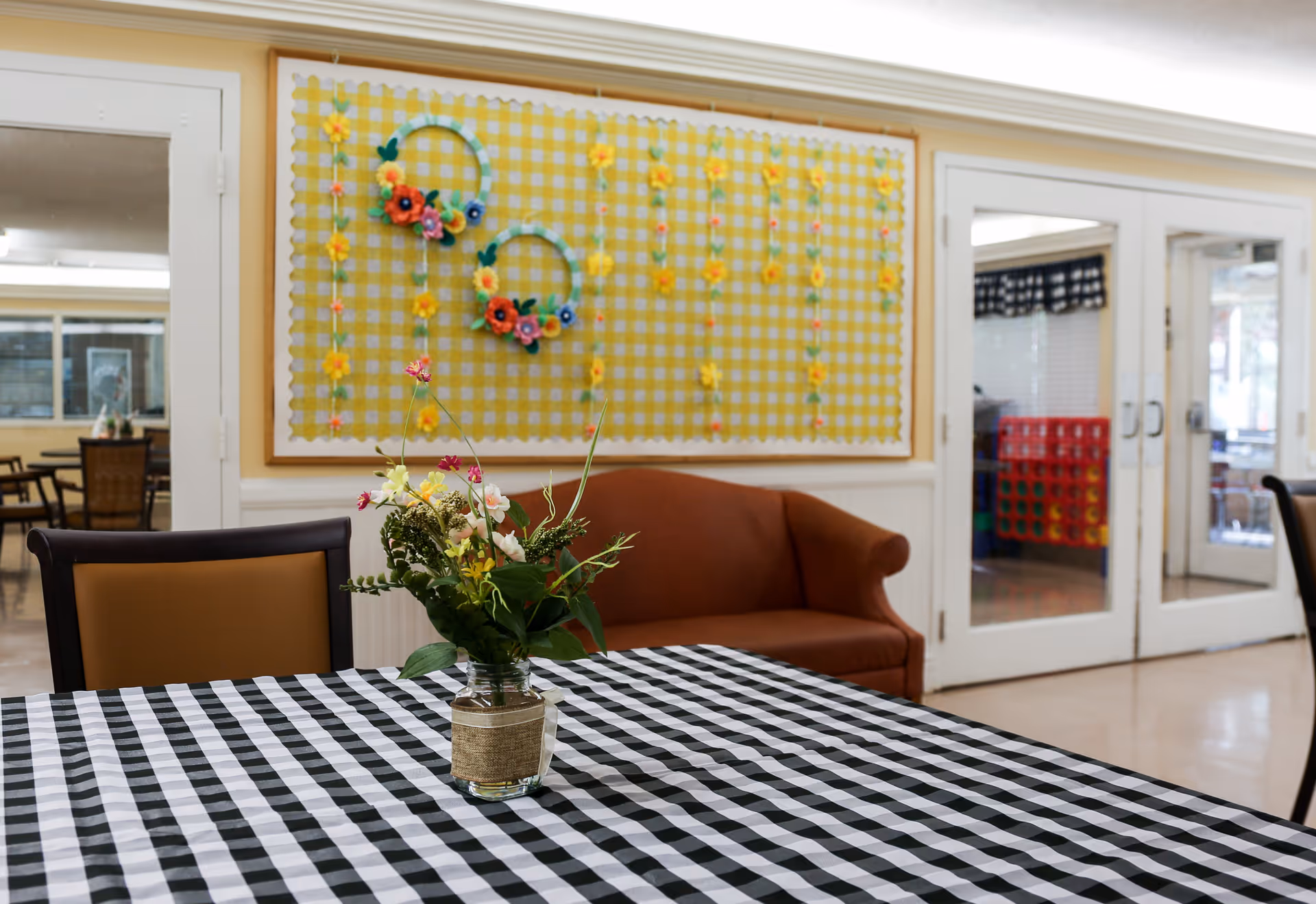 Interior view of a common area in a senior living facility featuring a table with a black and white checkered tablecloth and a small vase of flowers. Behind the table is a brown couch and a wall decorated with a yellow checkered fabric and colorful flower wreaths. Glass double doors lead to another room with visible chairs and a large Connect Four game.