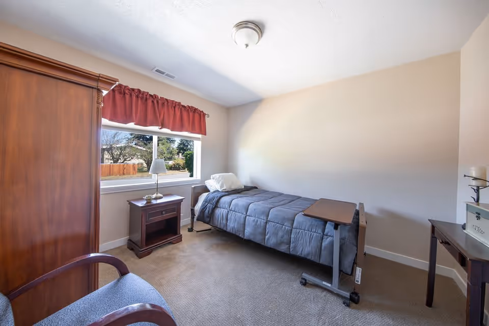 A small bedroom with a single bed covered in a gray comforter, a wooden nightstand with a lamp and decorative item, a wooden wardrobe, a chair with wooden arms, and a window with a red valance showing an outdoor view.