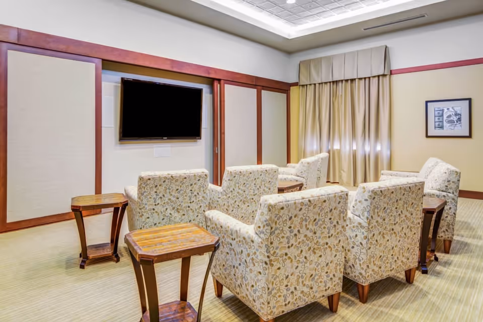 A small theater-style room with six patterned armchairs arranged in two rows facing a wall-mounted flat screen TV. The room has beige walls with wooden trim, a framed picture on the wall, and a window with beige curtains. There are small wooden side tables placed between the chairs.