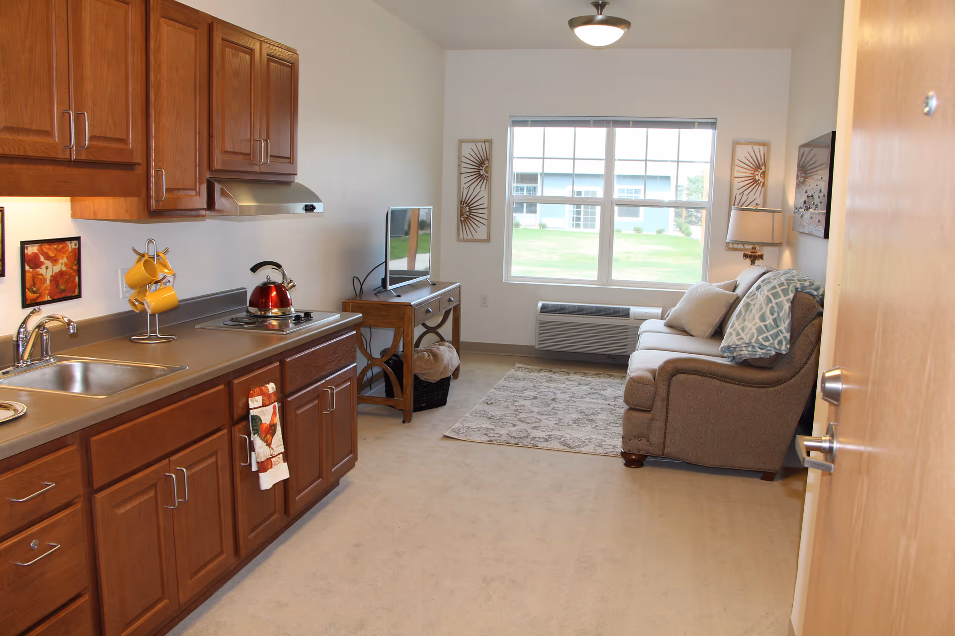 Interior view of a senior living facility apartment showing a small kitchen area with wooden cabinets, a sink, stovetop with a red kettle, and a rack holding yellow mugs. Adjacent to the kitchen is a living area with a brown sofa, a patterned rug, a wooden side table with a flat-screen TV, a lamp, and a large window overlooking a grassy outdoor area.