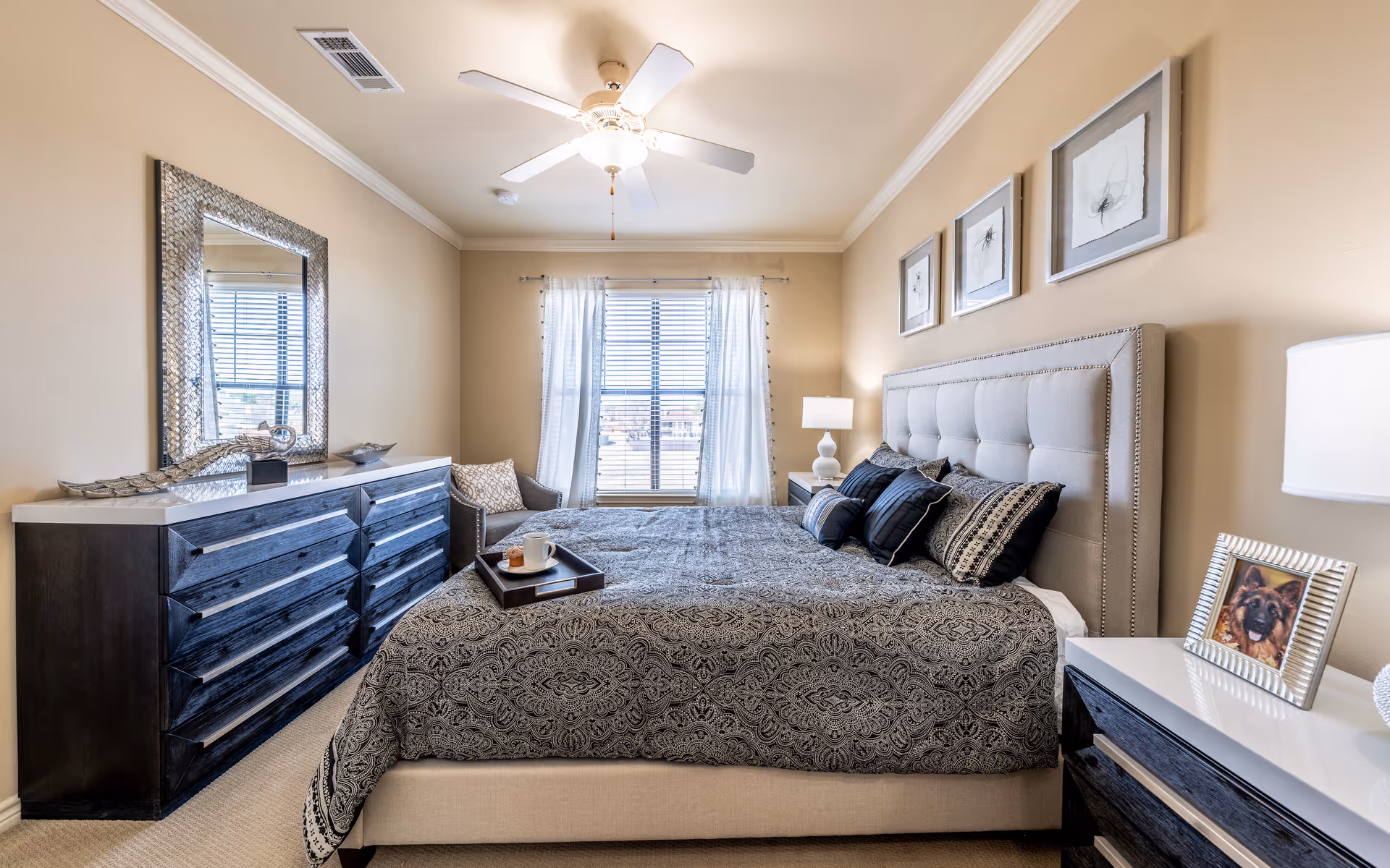 A well-lit bedroom with a large bed featuring a tufted headboard and patterned bedding. There are three framed artworks above the bed, a nightstand with a lamp and a framed photo of a dog, a dark wooden dresser with a decorative mirror above it, and a window with white curtains letting in natural light. A ceiling fan is mounted on the ceiling.