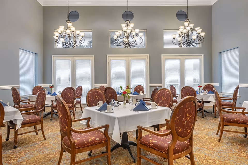 A bright dining room with multiple tables covered in white tablecloths, each set with blue folded napkins, cups, and glassware. The room features elegant wooden chairs with floral upholstery, large windows with blinds, and three ornate chandeliers hanging from the ceiling.