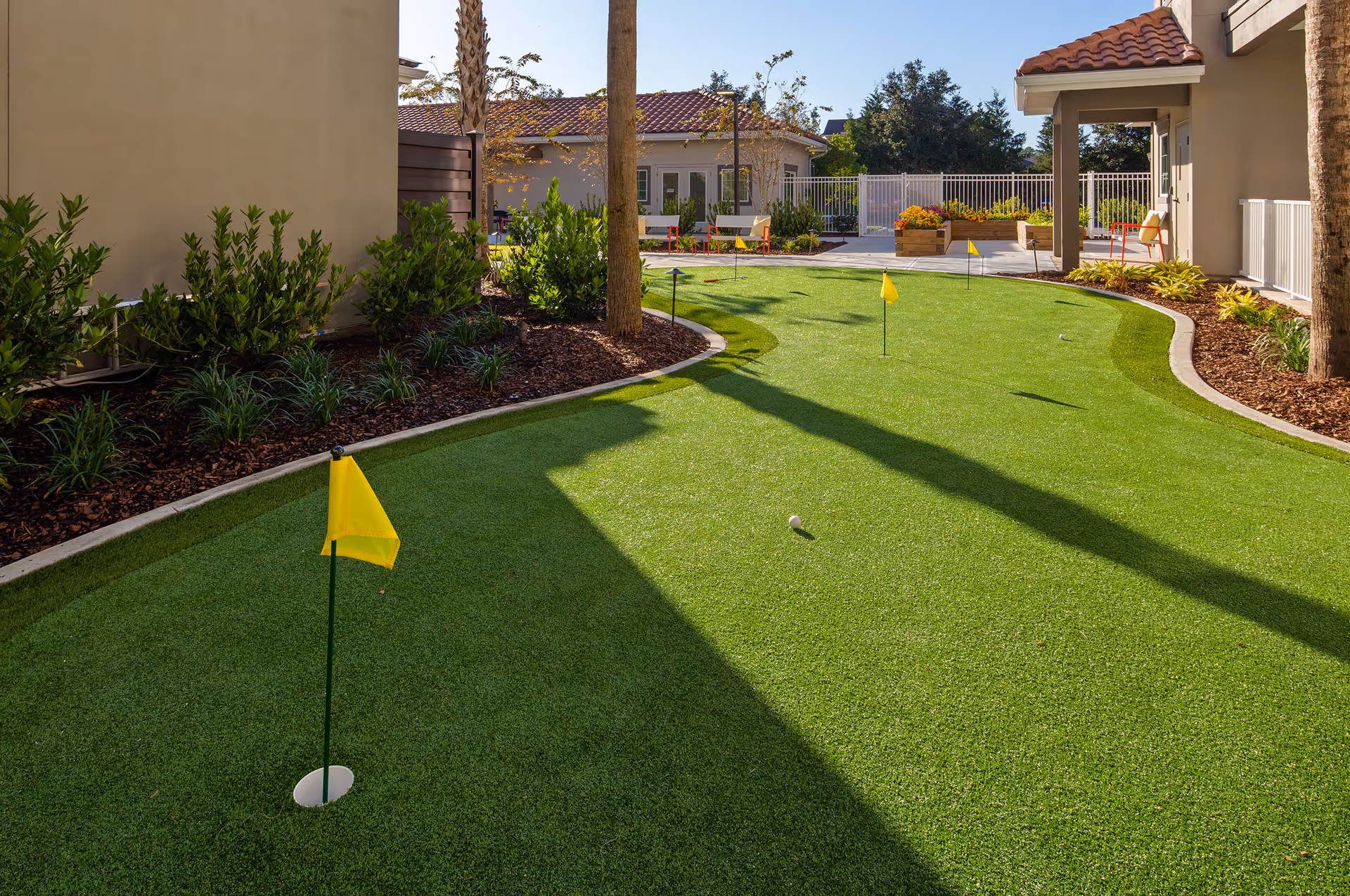 Outdoor putting green with several yellow flags on a sunny day, surrounded by plants, trees, and buildings with tiled roofs.