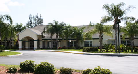 Exterior view of a single-story building with a covered entrance, surrounded by palm trees and landscaped greenery under a clear sky.