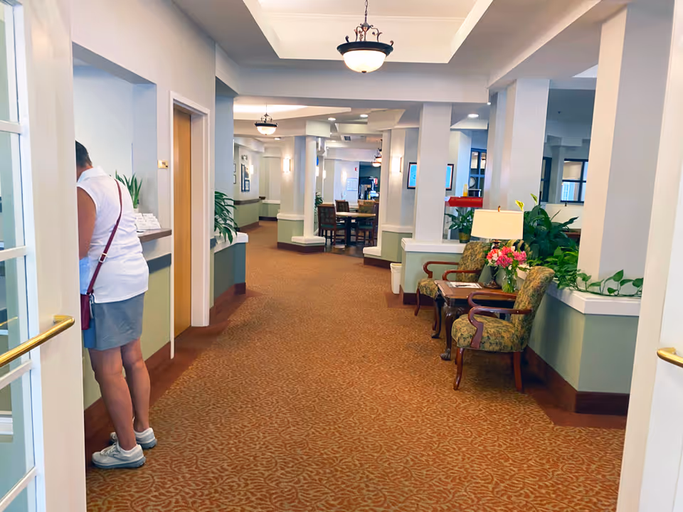 Interior hallway of a senior living facility with carpeted floor, white pillars, and soft lighting. There are two upholstered chairs with a small table and lamp between them on the right side, along with some plants. A woman in casual clothing is standing on the left side near a counter or window. In the background, there is a dining area with tables and chairs.