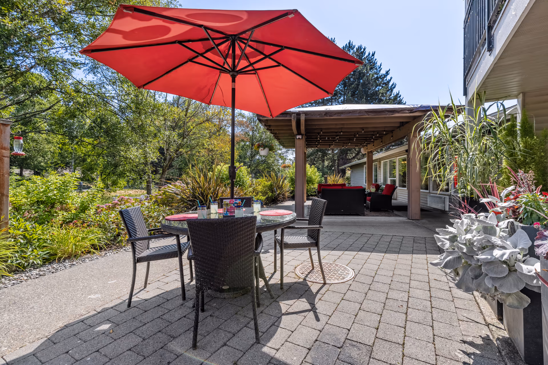 Outdoor patio area with a round table and four chairs under a large red umbrella. The patio is paved with stone tiles and surrounded by lush greenery and plants. In the background, there is a covered seating area with cushioned chairs and a table, attached to a building.
