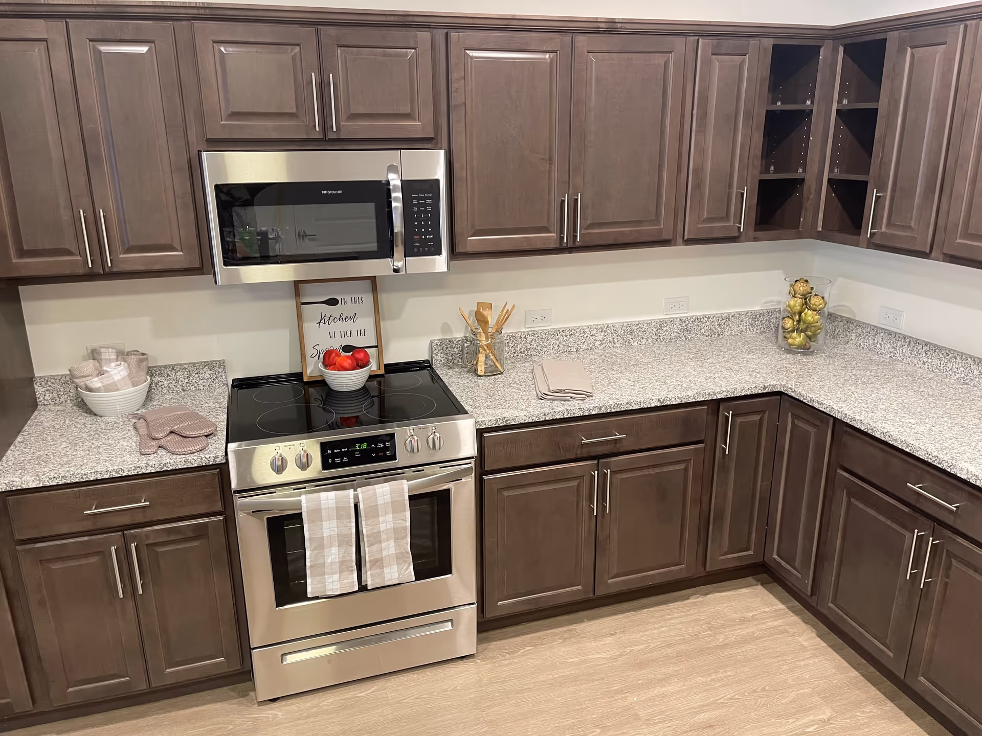 Modern kitchen with dark wood cabinets, granite countertops, a stainless steel electric stove with oven, and a microwave above it. The countertop has a bowl of red apples, a glass jar with artichokes, kitchen utensils in a holder, folded towels, and oven mitts.