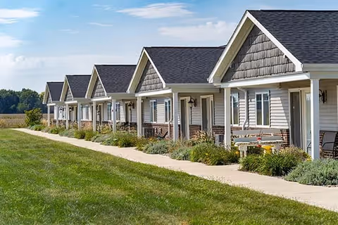 A row of single-story residential cottages with front porches, each featuring chairs and small tables, set alongside a paved walkway with green grass and landscaping in front under a partly cloudy sky.