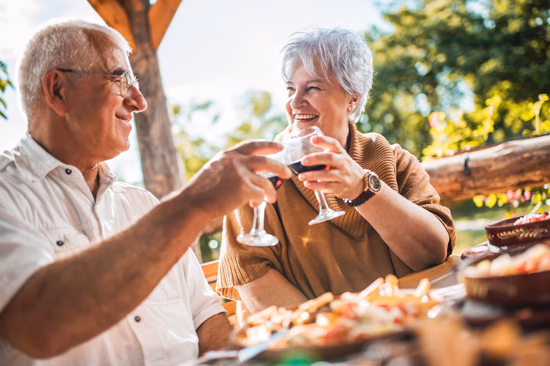 An elderly man and woman sitting outdoors at a wooden table, smiling and clinking glasses of red wine in a toast, with food on the table and greenery in the background.
