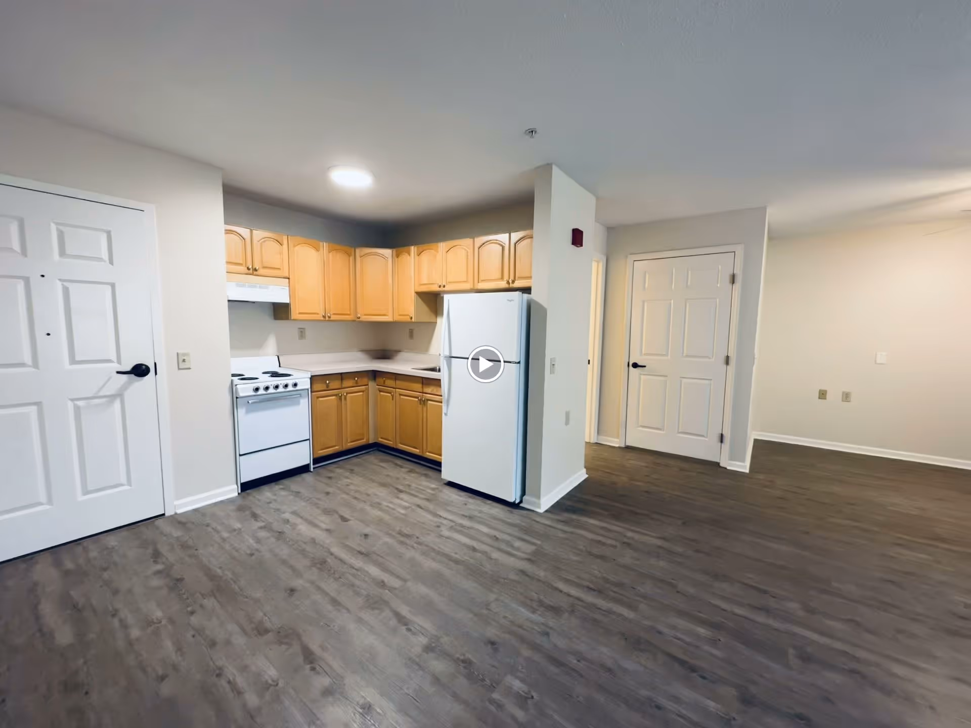 Open-plan apartment interior with a small kitchen featuring light wood cabinets, a white stove and refrigerator, and wood-look flooring.