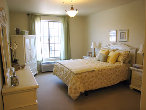 A bright and cozy bedroom with a large window covered by light green curtains. The room features a bed with a floral patterned bedspread and multiple pillows, two bedside tables with lamps, a white dresser, and a white armoire. The walls are light-colored and decorated with framed artwork.