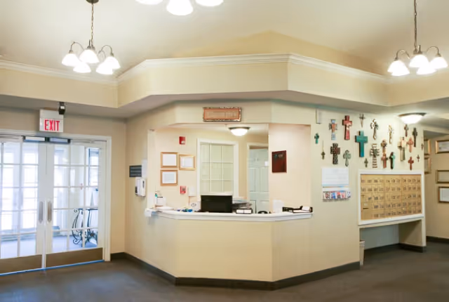 Reception area inside Trustwell Living at Terrebonne Place featuring a front desk with office supplies, a wall decorated with various crosses, a bulletin board with notices, and double glass doors with an exit sign above them.