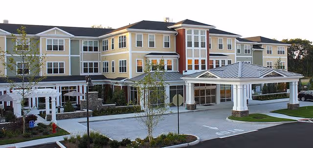 Exterior view of a large, multi-story senior living facility named One Wingate Way, featuring beige and red siding, multiple windows, a covered entrance with white columns, landscaped greenery, and a driveway with a stop sign.