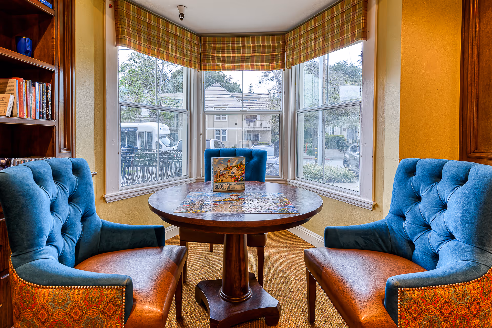 A cozy seating area in a senior living facility with a round wooden table and three blue upholstered chairs with brown leather seats. The table has a partially completed 300-piece puzzle on it. The room features large bay windows with plaid valances, allowing natural light to fill the space. A bookshelf filled with books is visible on the left side.