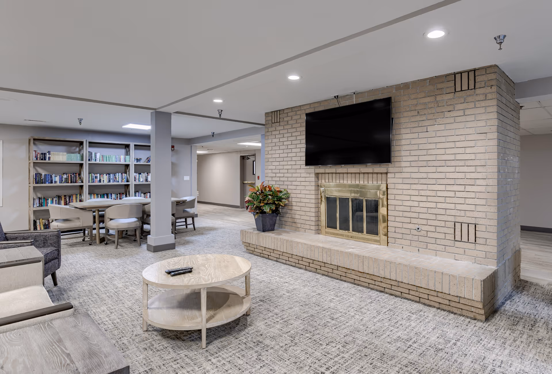 A spacious living room area with a beige brick fireplace and a mounted flat-screen TV above it. In front of the fireplace is a round wooden coffee table with a remote control on it. To the left, there are comfortable chairs and a wooden side table. In the background, there is a bookshelf filled with books and a table with chairs, suggesting a reading or activity area. The room has recessed lighting and neutral-colored walls and carpet.