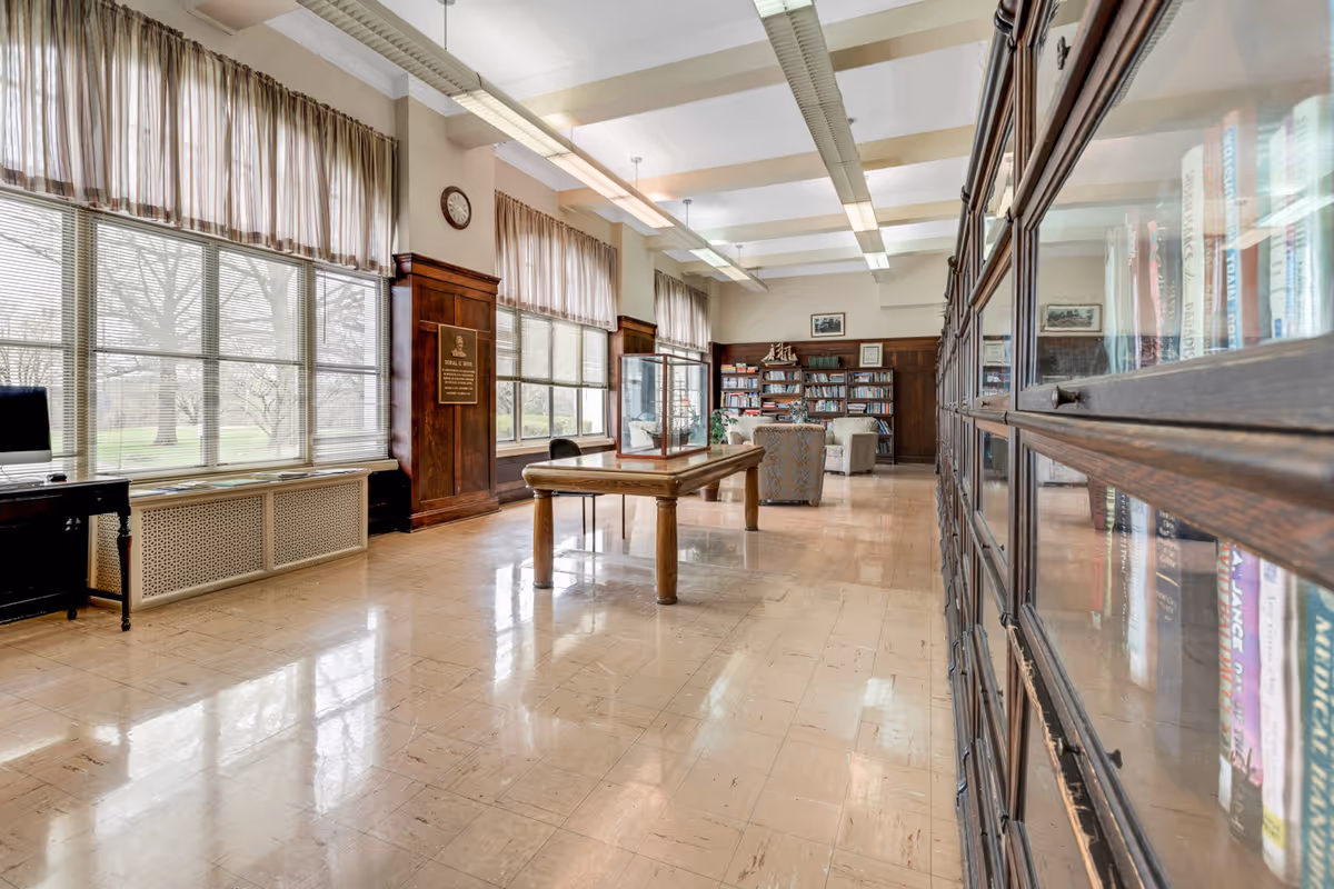 A spacious, well-lit library or reading room with large windows covered by sheer curtains, wooden bookshelves filled with books, a wooden table in the center, and comfortable armchairs near the back wall. The room has a polished tile floor and a high ceiling with exposed beams and fluorescent lighting.