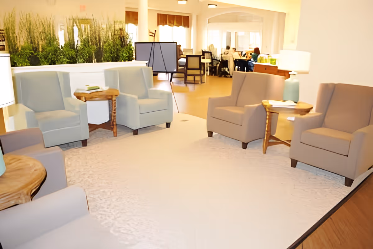 Bright communal lounge with several armchairs and side tables arranged on a light rug, with plants and a dining area visible in the background.