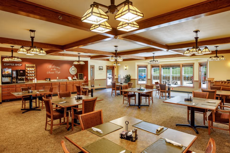 Well-lit dining room with multiple tables and chairs, a coffee station along the back wall, and large windows letting in natural light.